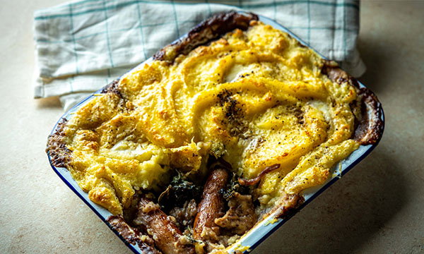 Baked casserole with golden mashed potato swirls on top, crispy dark edges, set on a light surface with a green checked cloth underneath. Comforting and rustic.