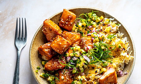 A bowl of glazed salmon cubes with sesame seeds and colourful diced vegetables on rice, garnished with spring onions. A fork lies beside it, creating a fresh, vibrant look.