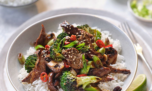Stir-fry beef with broccoli, mushrooms, and red chilli on rice, garnished with sesame seeds and green onions in a grey bowl, conveying a savoury and vibrant presentation.