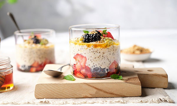A glass jar filled with layered overnight oats topped with strawberries, blackberries, and mint sits on a wooden board. Honey and oats are in the background.