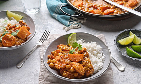 A bowl of curry with chickpeas and sweet potatoes is served alongside white rice, garnished with a lime wedge and coriander, evoking a hearty, warm meal.