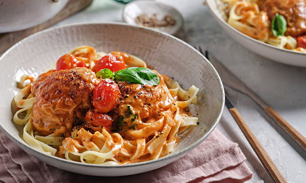 A bowl of creamy pasta topped with two chicken thighs, cherry tomatoes, and fresh basil. The dish is garnished with pepper, set on a pink napkin, creating a cosy vibe.