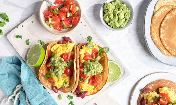 Two breakfast tacos with scrambled eggs, tomatoes, and guacamole on a board, surrounded by lime halves, salsa, and a side of corn tortillas. Bright and fresh ambiance.