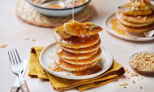 Fluffy pancake stack topped with caramelised bananas and drizzled caramel sauce. Fork and knife on yellow napkin beside plate. Warm, inviting setting.
