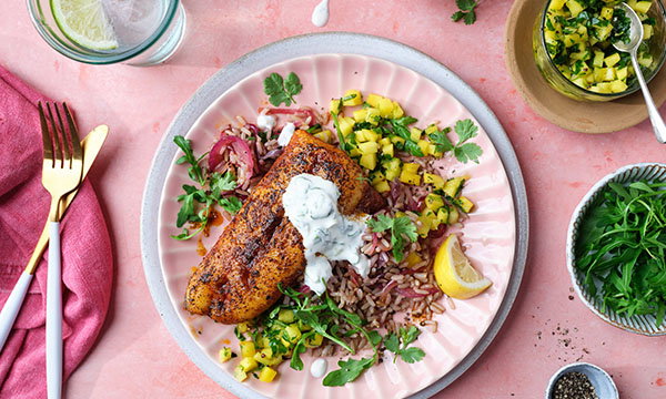 Grilled fish with creamy sauce on a pink plate, served with wild rice, pineapple salsa, rocket, and lemon wedge. Cutlery and a drink are nearby.