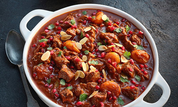 White dish with a rich beef stew, featuring pomegranate seeds and almond slivers. Garnished with cilantro. Warm, hearty, and inviting.