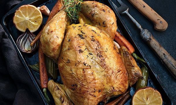 Golden roast chicken on a tray with thyme garnish, surrounded by roasted carrots, celery, onions, and lemon halves. Rustic cooking tools nearby.