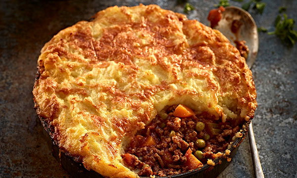 Golden-brown shepherd's pie with a crust of mashed potatoes. A serving is missing, revealing minced meat, peas, and carrots. A spoon lies beside the dish.