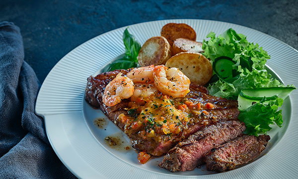 A plate of steak topped with grilled prawns and sauce, accompanied by roasted potatoes and a fresh salad of lettuce and cucumber slices.