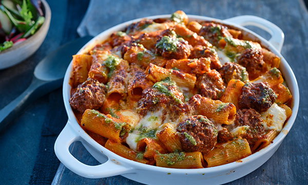 A baked pasta dish in a white casserole; includes rigatoni, meatballs, melted cheese, and green herbs. The setting is a rustic wooden table.