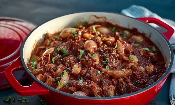 A red pot filled with rich, savoury beef stew, featuring chunks of beef, mushrooms, onions, and herbs, emits a hearty and comforting aroma.