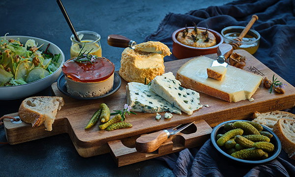 Cheese board with assorted cheeses, p&acirc;t&eacute; with a reddish topping, cornichons, fresh bread, and salad. The scene is warm and rustic.