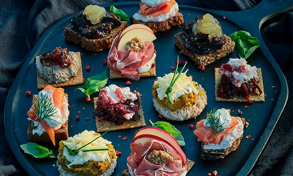 A platter of gourmet canapés features various open-faced sandwiches with toppings like smoked salmon, prosciutto, apple slices, and herbs, set on rustic bread.