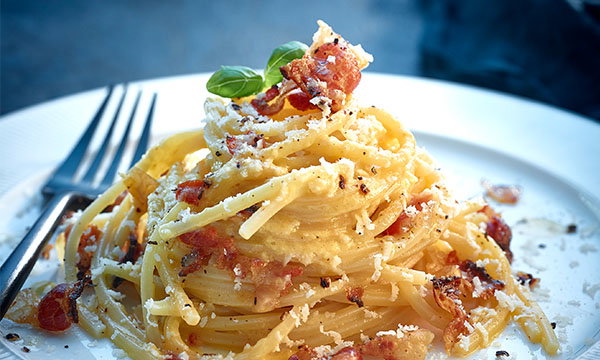 A plate of spaghetti carbonara is topped with crispy bacon, grated cheese, and fresh basil leaves, evoking a rustic and appetising feel.