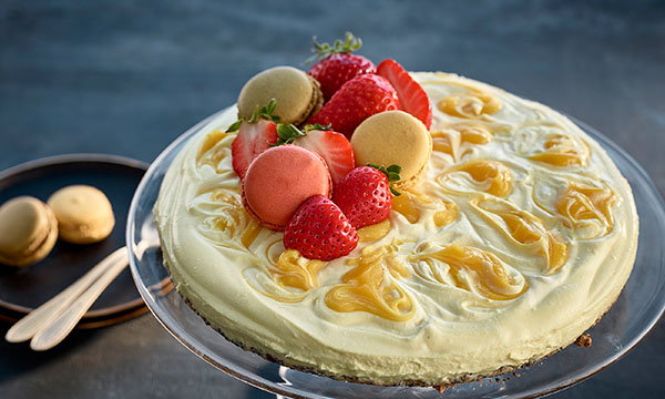 Cheesecake topped with strawberries and assorted macarons, elegantly swirled with mango puree on a glass stand. A plate with macarons nearby.