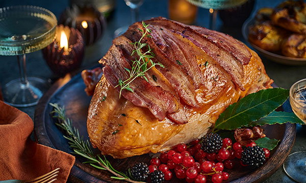 Roasted turkey breast topped with crispy bacon and herbs on a platter, surrounded by red currants, blackberries, and a leaf garnish. Cosy, festive ambiance.