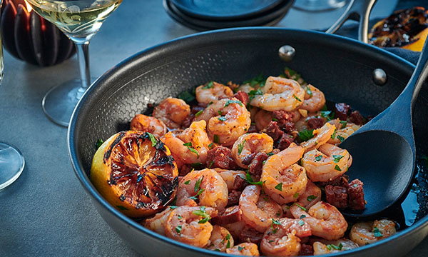 A dish of saut&eacute;ed prawns and chorizo garnished with parsley in a black pan, with charred lemon slices. Two wine glasses sit in the background.