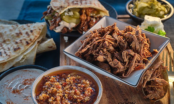 A spread of shredded meat in a bowl, with a barbecue sauce dish, pita bread, and a sandwich topped with pickles. Rustic and appetising presentation.