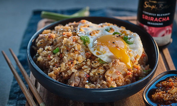 A bowl of fried rice topped with a runny sunny-side-up egg and green onions, served with a bottle of Sriracha sauce and chopsticks on a wooden board.