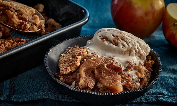 A bowl of warm apple crisp topped with melting ice cream sits on a blue cloth. A baking dish and whole apples are in the background.