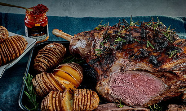 Roasted leg of lamb garnished with herbs and charred bits, surrounded by hasselback potatoes. A jar of spicy spread is open nearby on a blue cloth.