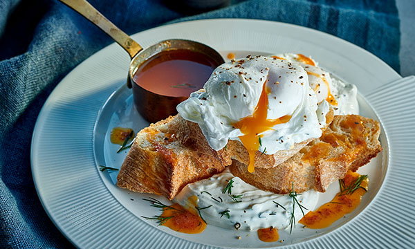 A rustic plate with poached eggs on crusty bread, drizzled with sauce and herbs. The yolk seeps onto the bread, next to a small cup of sauce.