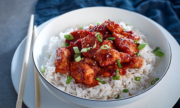 A white bowl filled with white rice topped with glazed, red-colored chicken pieces and garnished with chopped green onions. Chopsticks rest beside the bowl.