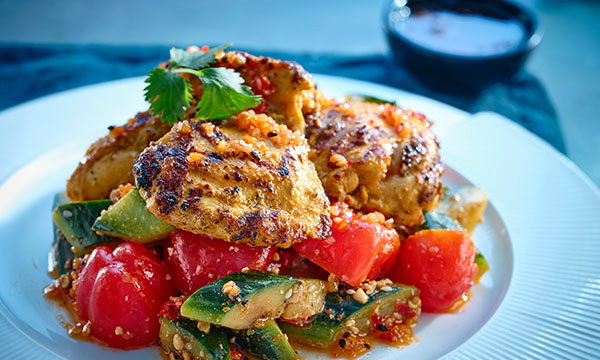 Grilled chicken on a bed of roasted cherry tomatoes and peppers, garnished with coriander, on a white plate; a small bowl of sauce in the background.