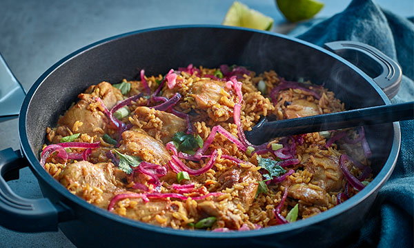 A black skillet filled with seasoned chicken pieces, vibrant red onions, and rice garnished with fresh herbs, emits comforting steam on a table.