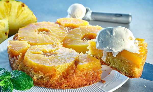 Pineapple upside-down cake with caramelised pineapple rings on top, a slice being served with whipped cream, and mint leaves beside it on a white plate.