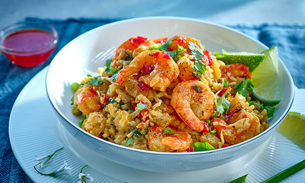 A vibrant dish of prawn fried rice garnished with coriander, green onions, and a slice of lime. A small bowl of red dipping sauce sits beside it.