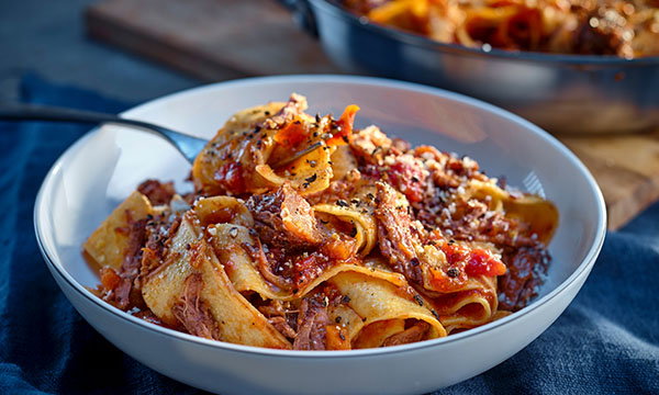 A white bowl filled with wide pappardelle pasta and rich meat sauce, garnished with grated cheese and black pepper, on a blue cloth. Warm, inviting, and comforting.
