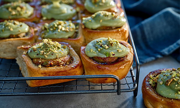 Freshly baked cinnamon rolls topped with pistachio green icing and chopped pistachios sit on a cooling rack. A cozy, inviting atmosphere is conveyed.