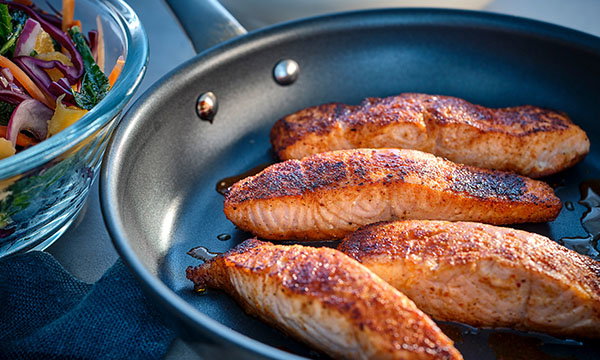 Three well-seasoned salmon fillets searing in a dark non-stick pan, alongside a fresh salad with vibrant greens and shredded carrots, creating a warm, appetising scene.