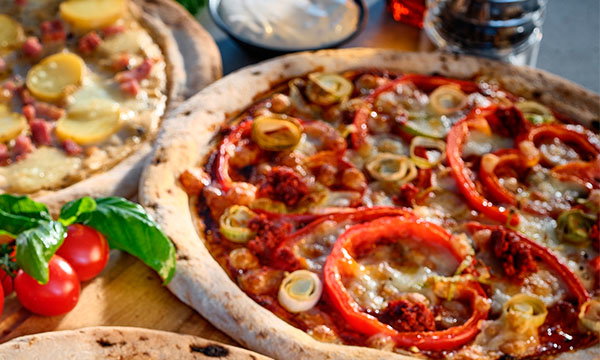 A close-up of two pizzas on a wooden table. One pizza features red peppers and rings of leeks. Fresh tomatoes and basil add a vibrant touch.