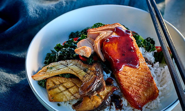 Salmon rice bowl topped with glazed salmon, saut&eacute;ed mushrooms, leafy greens and rice, served with chopsticks