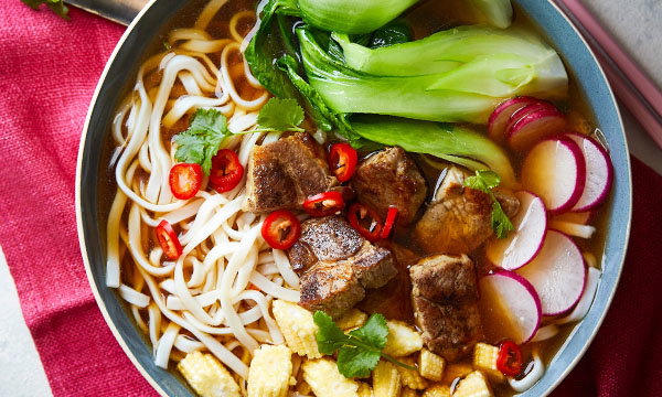 A bowl of noodle soup with pak choi beef chunks, sliced radishes, baby corn, and red chilies, garnished with coriander, on a red cloth background.
