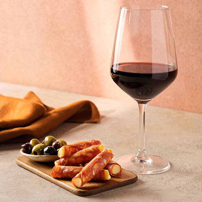 Glass of red wine with a wooden board of rolled chorizo cheese sticks and mixed olives on a beige surface.