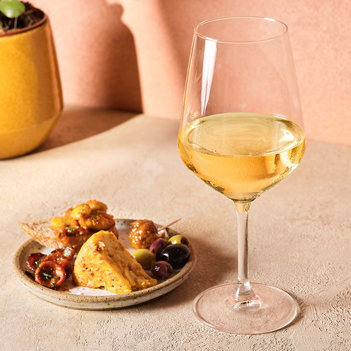 Glass of white wine beside a small plate of assorted appetizers including olives, cheese, and skewered items on a textured surface.
